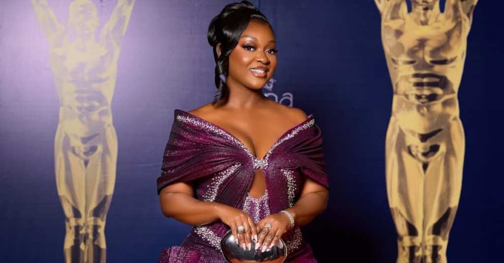 Jackie Appiah wearing a sculptural metallic magenta-purple gown with origami-inspired pleats and rhinestone detailing at the 2026 Ghana Movie Awards red carpet in Accra.