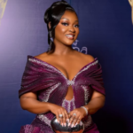 Jackie Appiah wearing a sculptural metallic magenta-purple gown with origami-inspired pleats and rhinestone detailing at the 2026 Ghana Movie Awards red carpet in Accra.