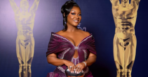 Jackie Appiah wearing a sculptural metallic magenta-purple gown with origami-inspired pleats and rhinestone detailing at the 2026 Ghana Movie Awards red carpet in Accra.